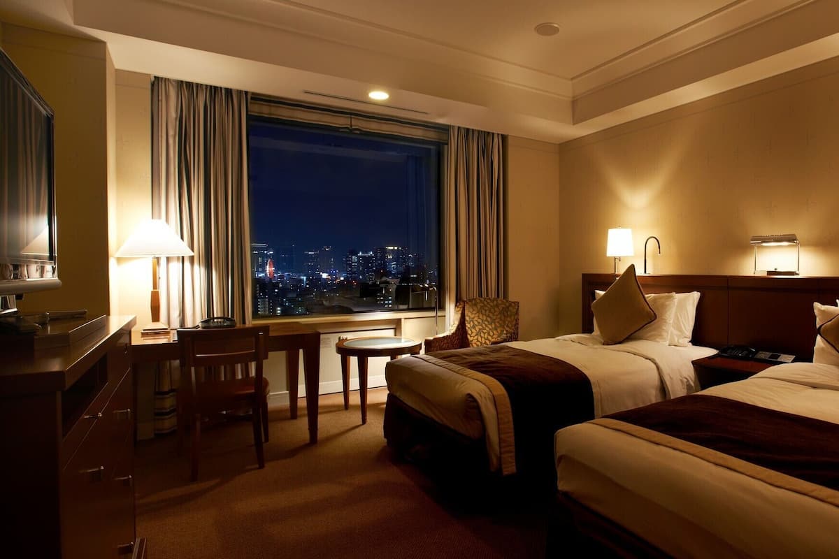 A hotel room with two beds, a desk, a chair, a lamp, and a view of the city at night.