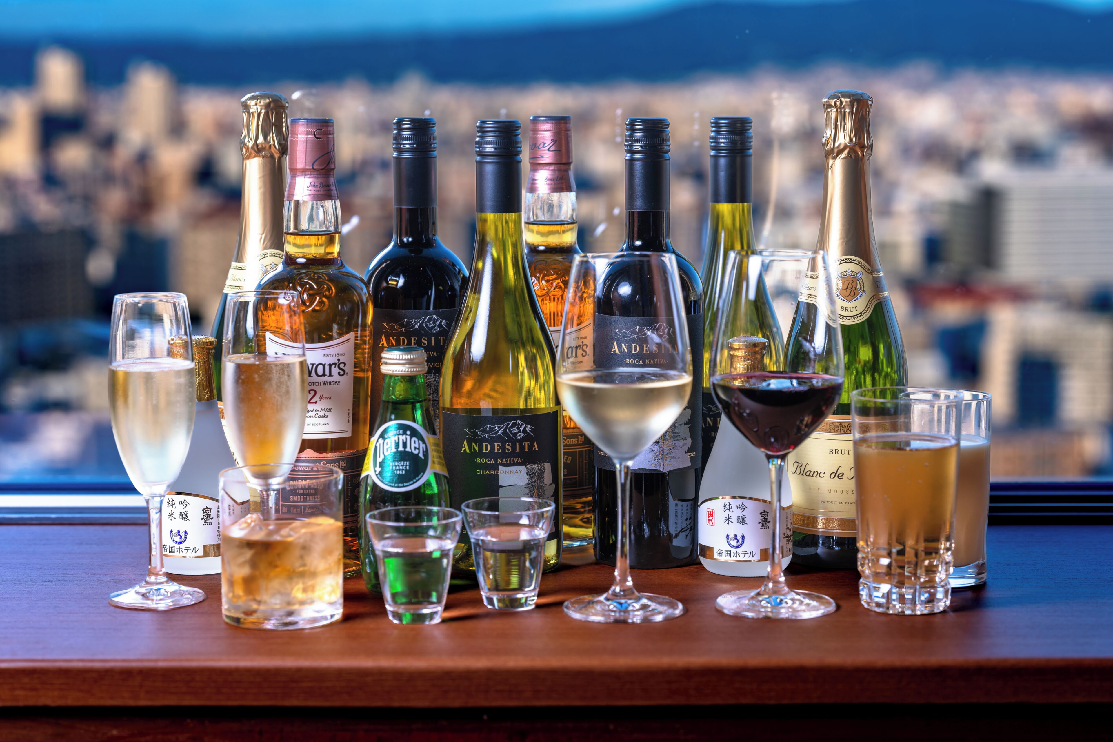 Variedad de botellas y copas sobre una barra con vistas a un paisaje urbano al fondo.