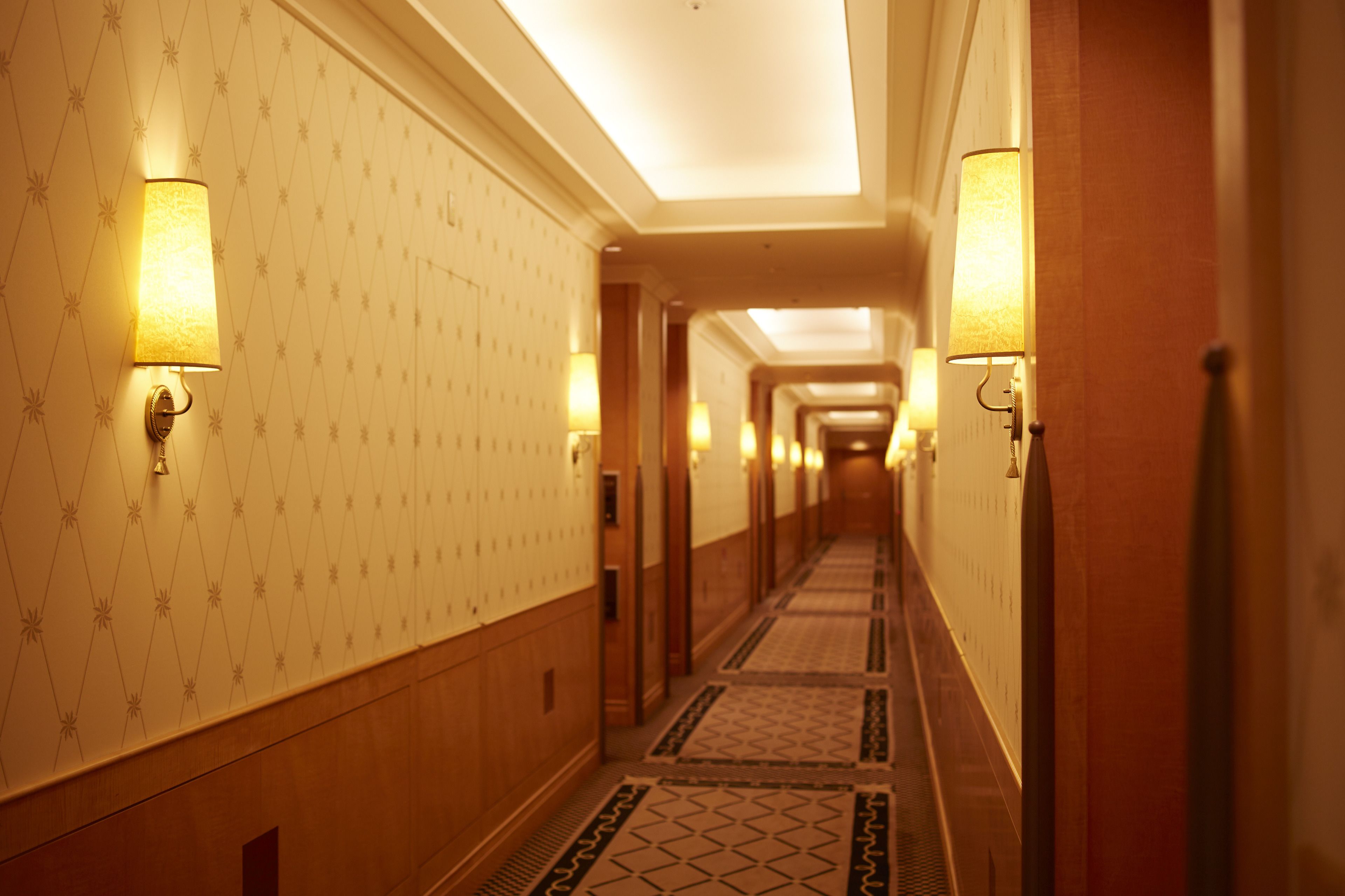 A hotel hallway with patterned wallpaper, wall-mounted lamps, and a tiled floor.