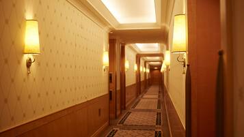 A hotel hallway with patterned wallpaper, wall-mounted lamps, and a tiled floor.