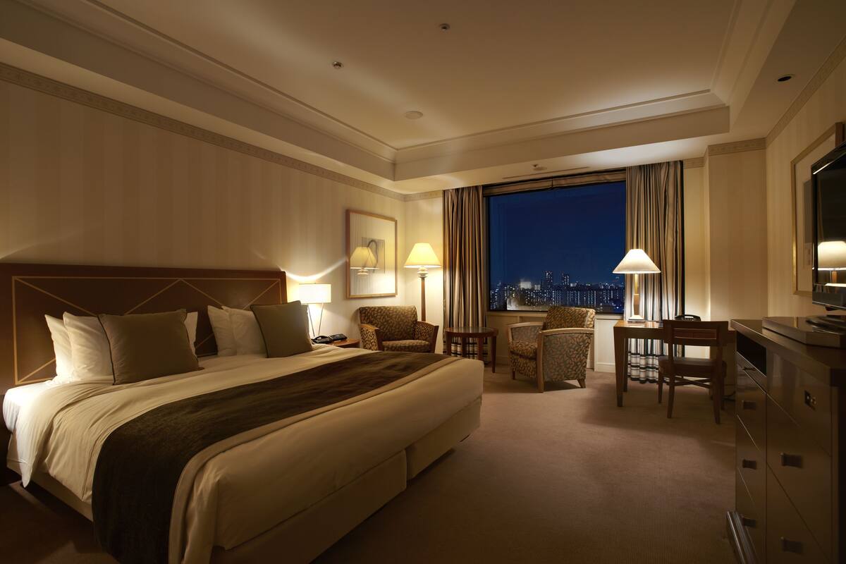 A hotel room with a large bed, two armchairs, a desk, and a view of the city at night.