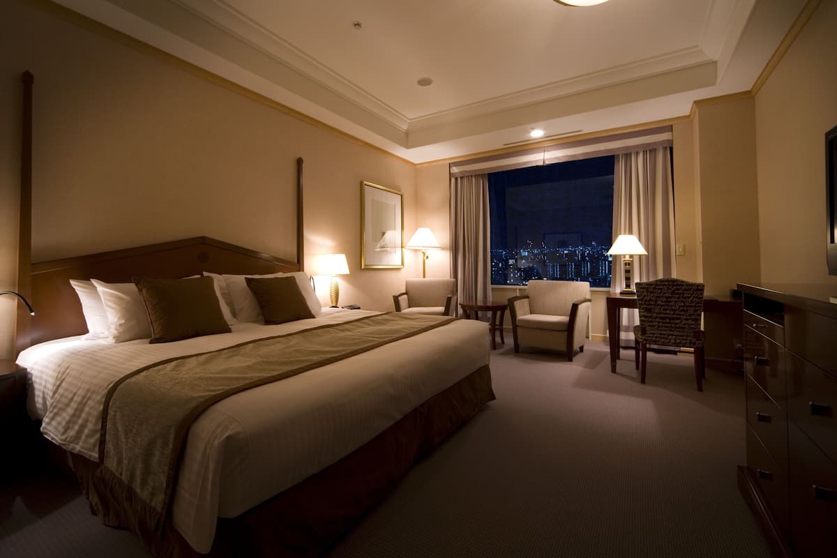 A hotel room with a large bed, two chairs, a desk, and a view of a cityscape at night.
