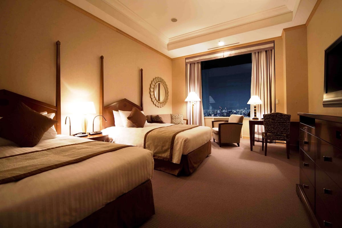 A hotel room with two beds, a desk, a chair, a TV, and a large window with a city view.
