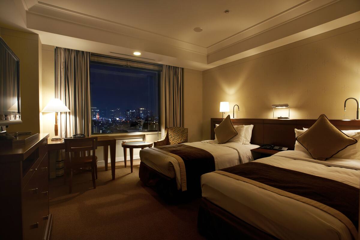 A hotel room with two beds, a desk with a chair, a small table, and a view of the city at night.