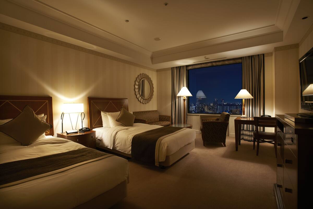 A hotel room with two beds, a desk, a chair, and a view of the cityscape at night.
