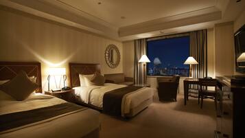A hotel room with two beds, a desk, and a view of the cityscape at night.