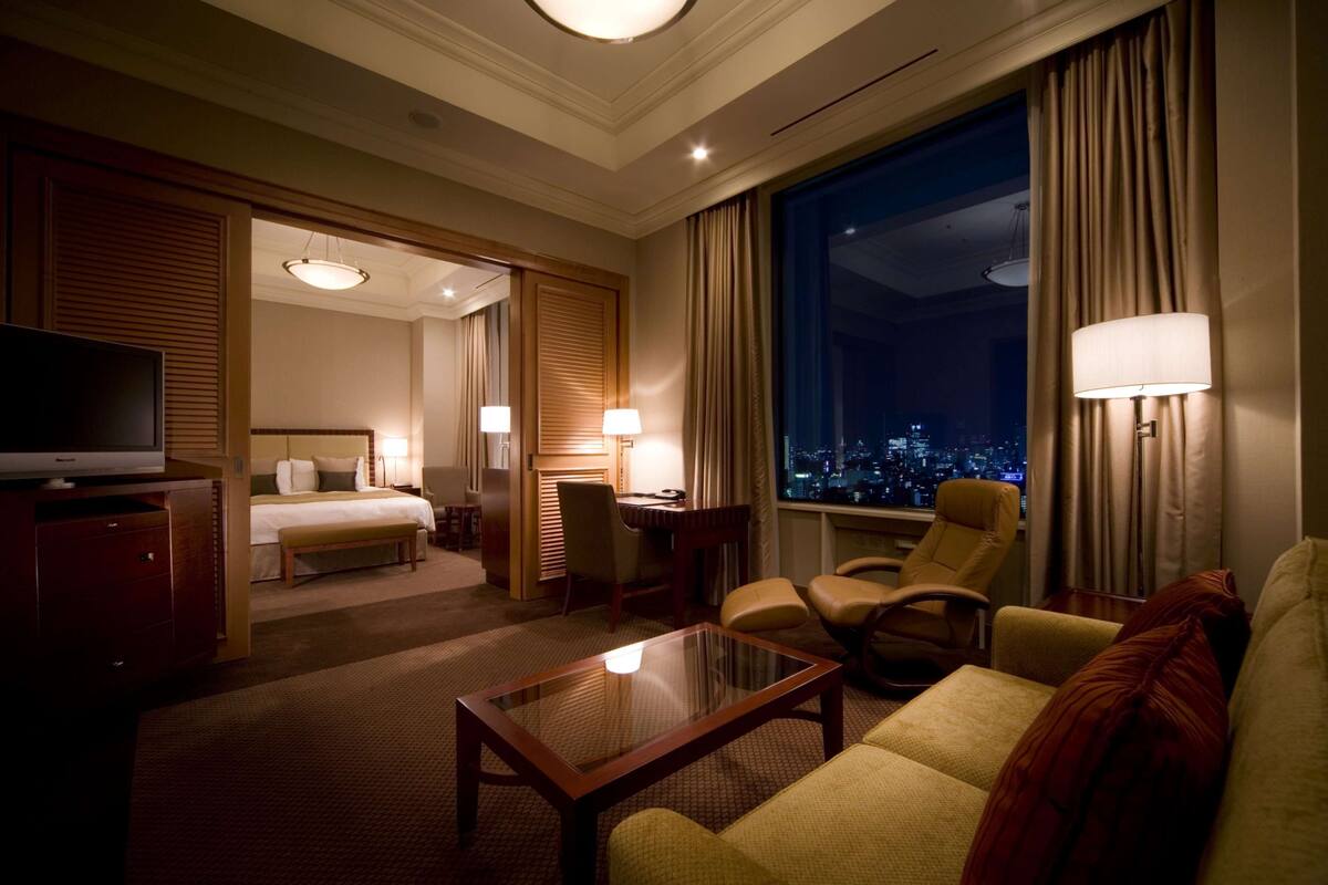 A hotel room with a bed, a TV, a desk, a chair, a sofa, and a window with a city view.