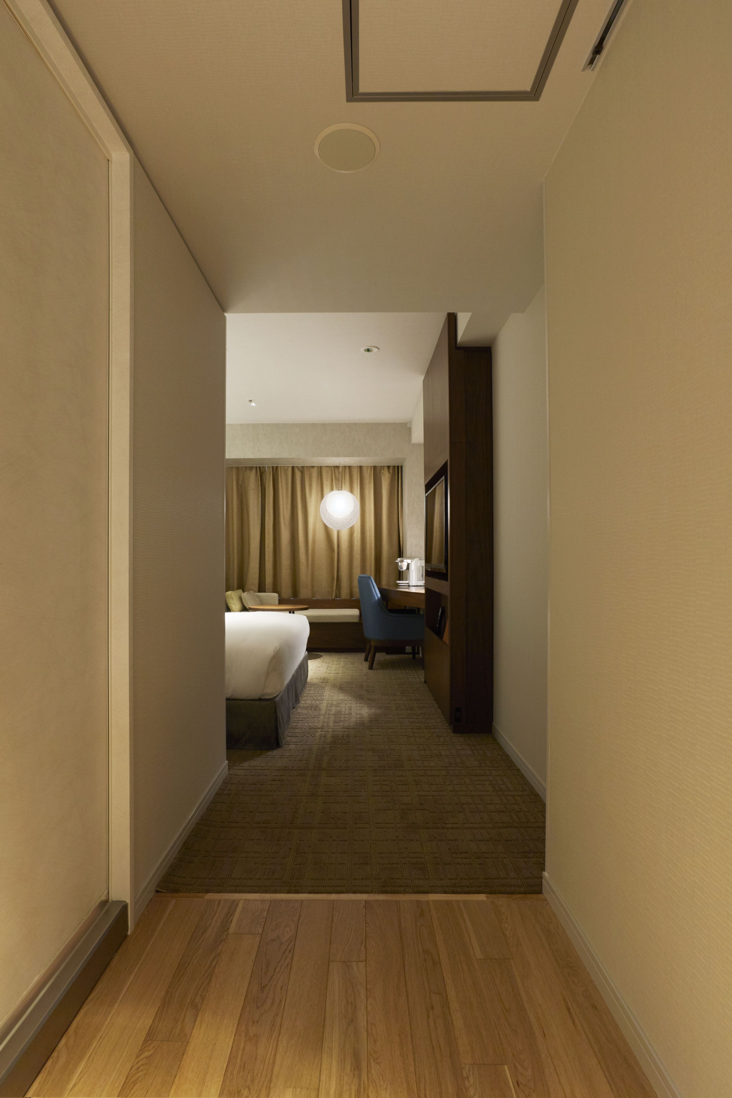 A hotel hallway leading to a bedroom with a bed, a desk, and a chair.
