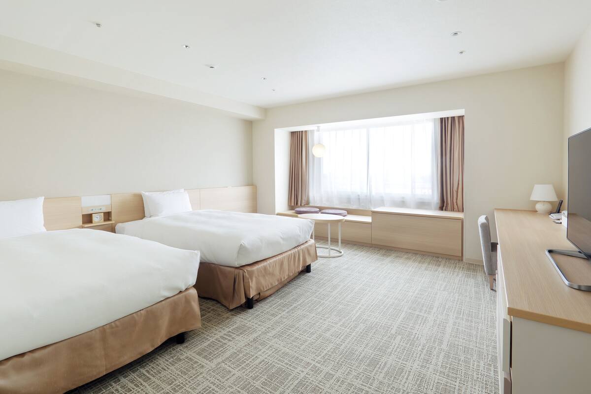 Ein Hotelzimmer mit zwei Betten, einem Schreibtisch, einem Fernseher und einem Fenster mit Vorhängen.