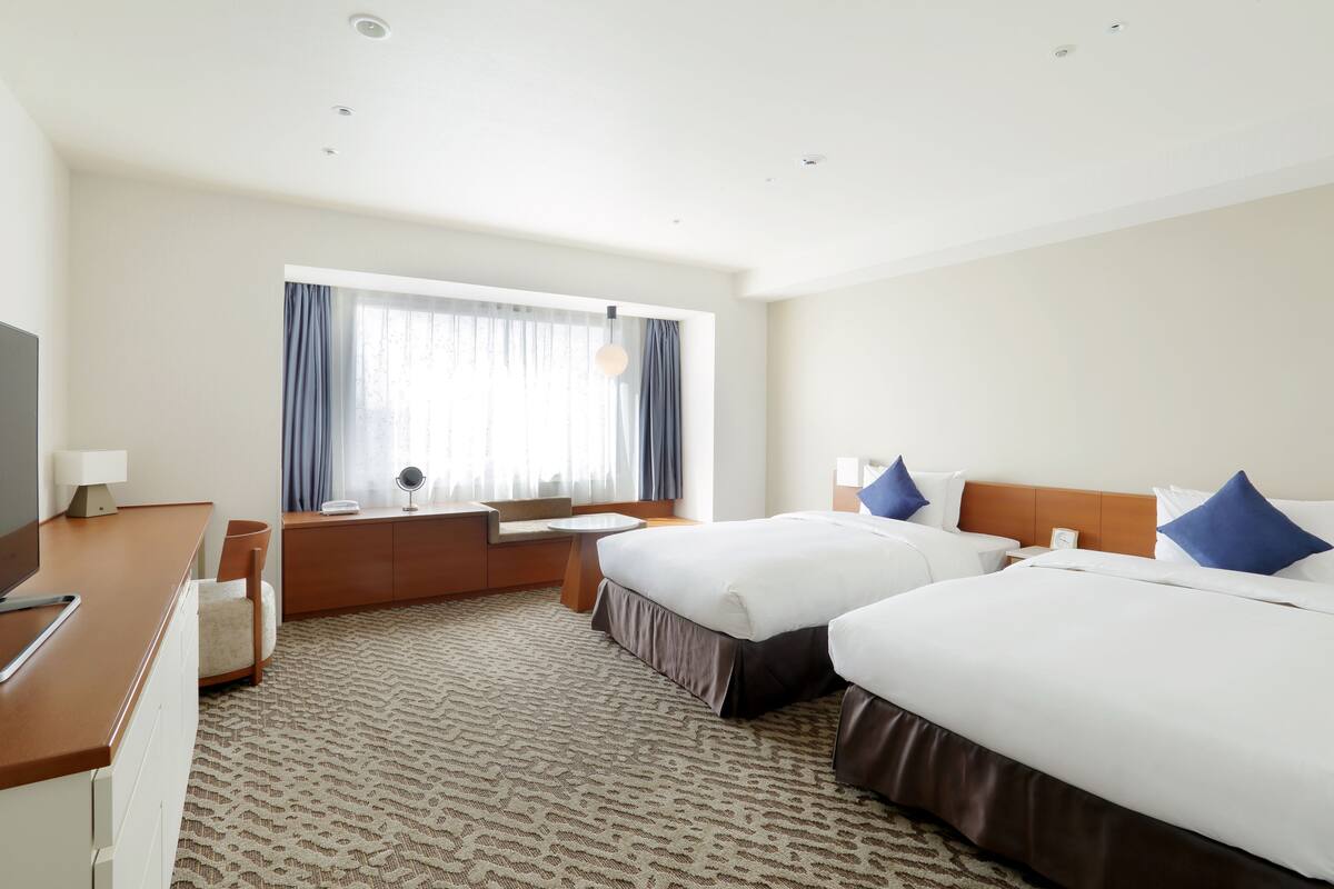 Ein Hotelzimmer mit zwei Betten, einem Schreibtisch, einem Stuhl und einem großen Fenster mit Vorhängen.