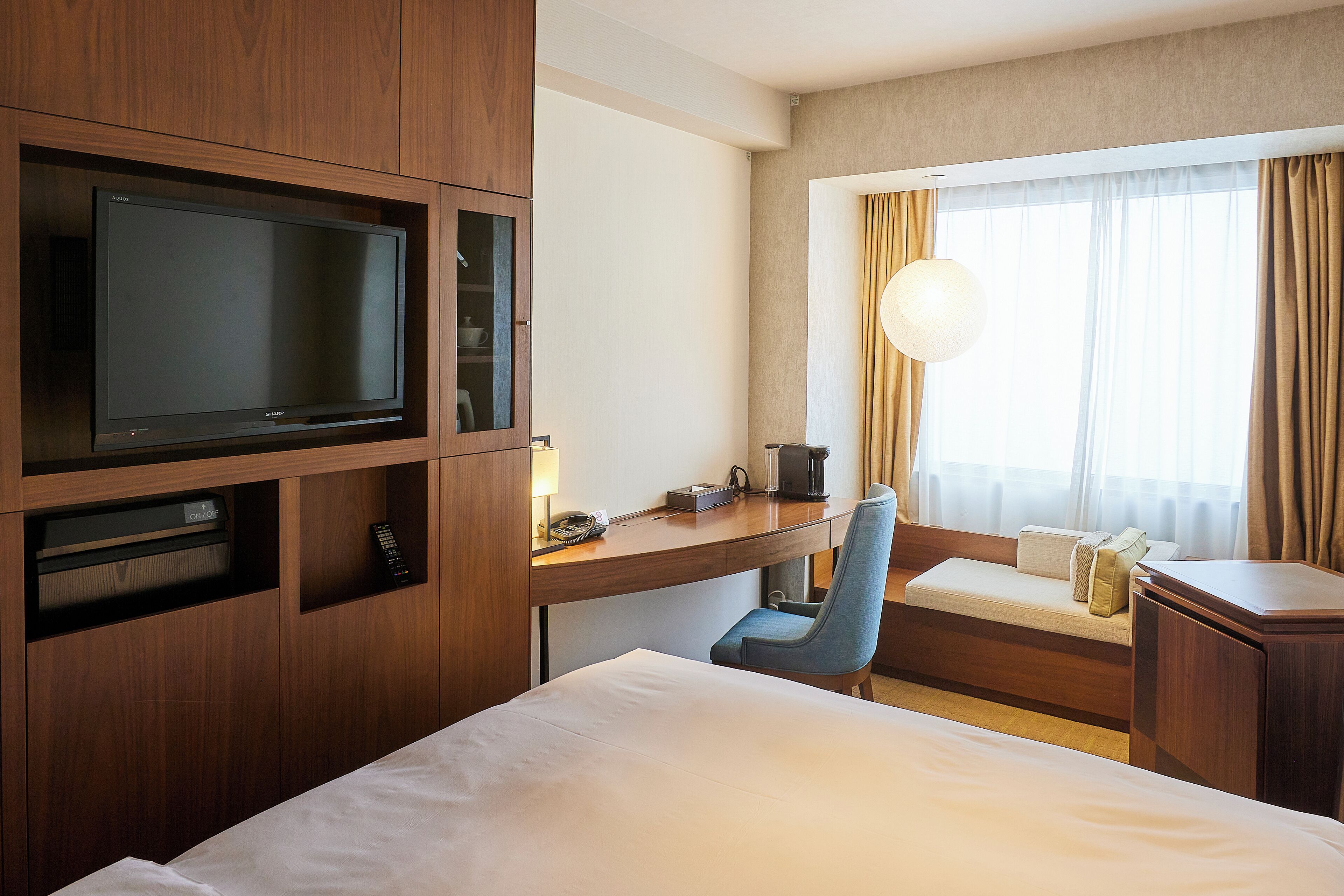 A hotel room with a bed, a desk with a television, a chair, and a window with curtains.