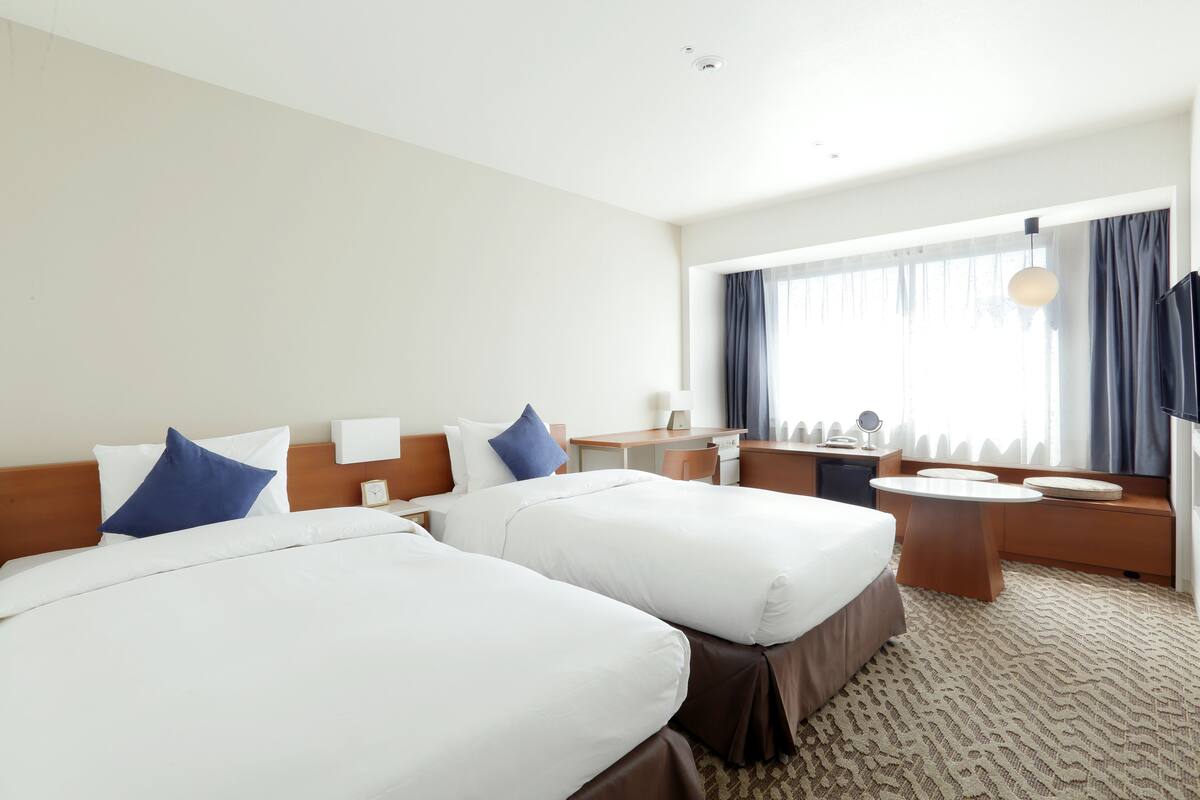 Ein Hotelzimmer mit zwei Betten, einem Schreibtisch, einem Stuhl, einem kleinen Tisch und einem großen Fenster mit Vorhängen.
