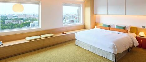 A modern hotel room with a large bed, built-in seating area, and large windows offering city views.