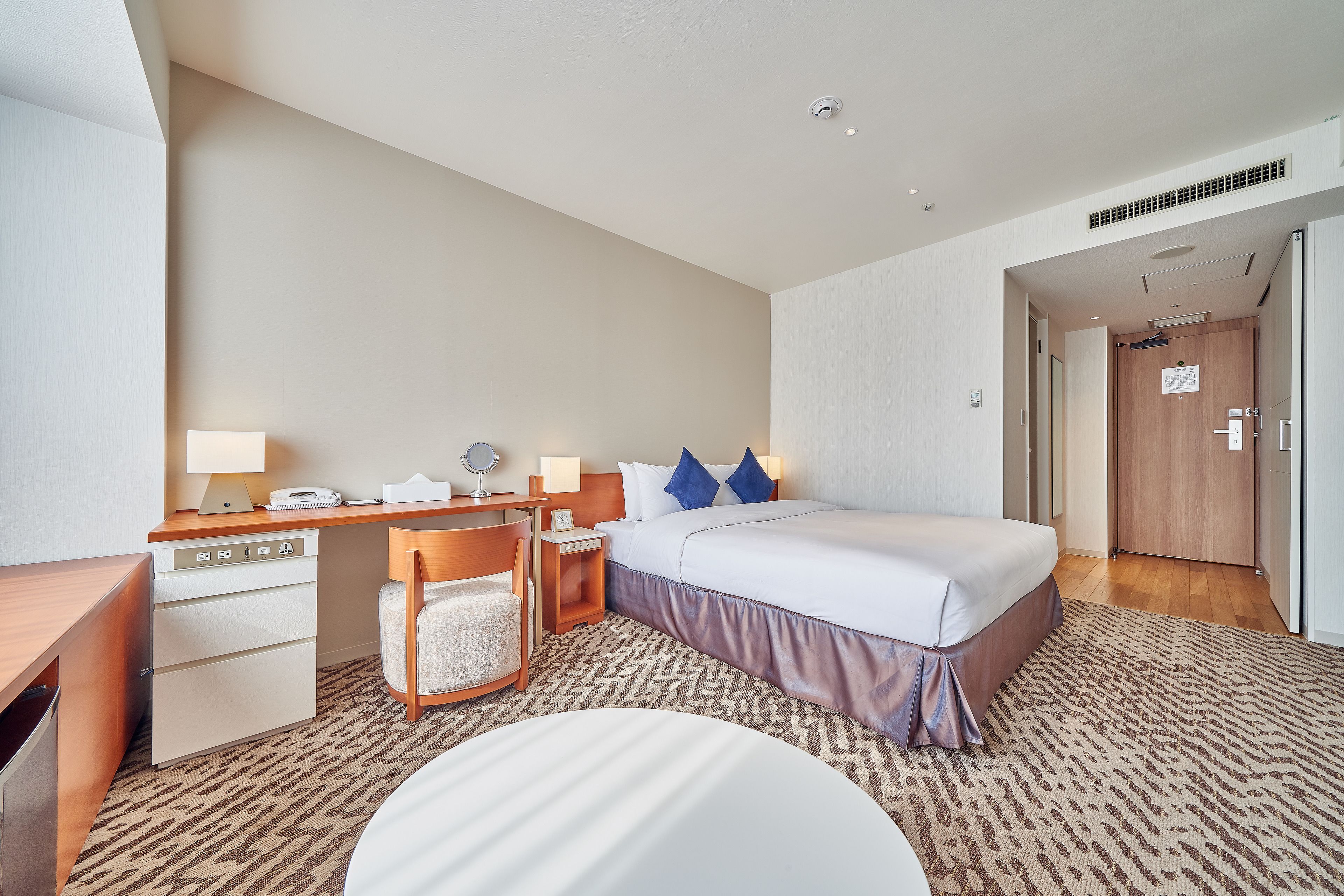A hotel room with a bed, desk, chair, and a small table with a lamp.