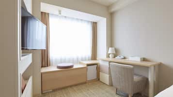 A hotel room with a bed, a desk with a chair, a small refrigerator, a TV, and a window with curtains.