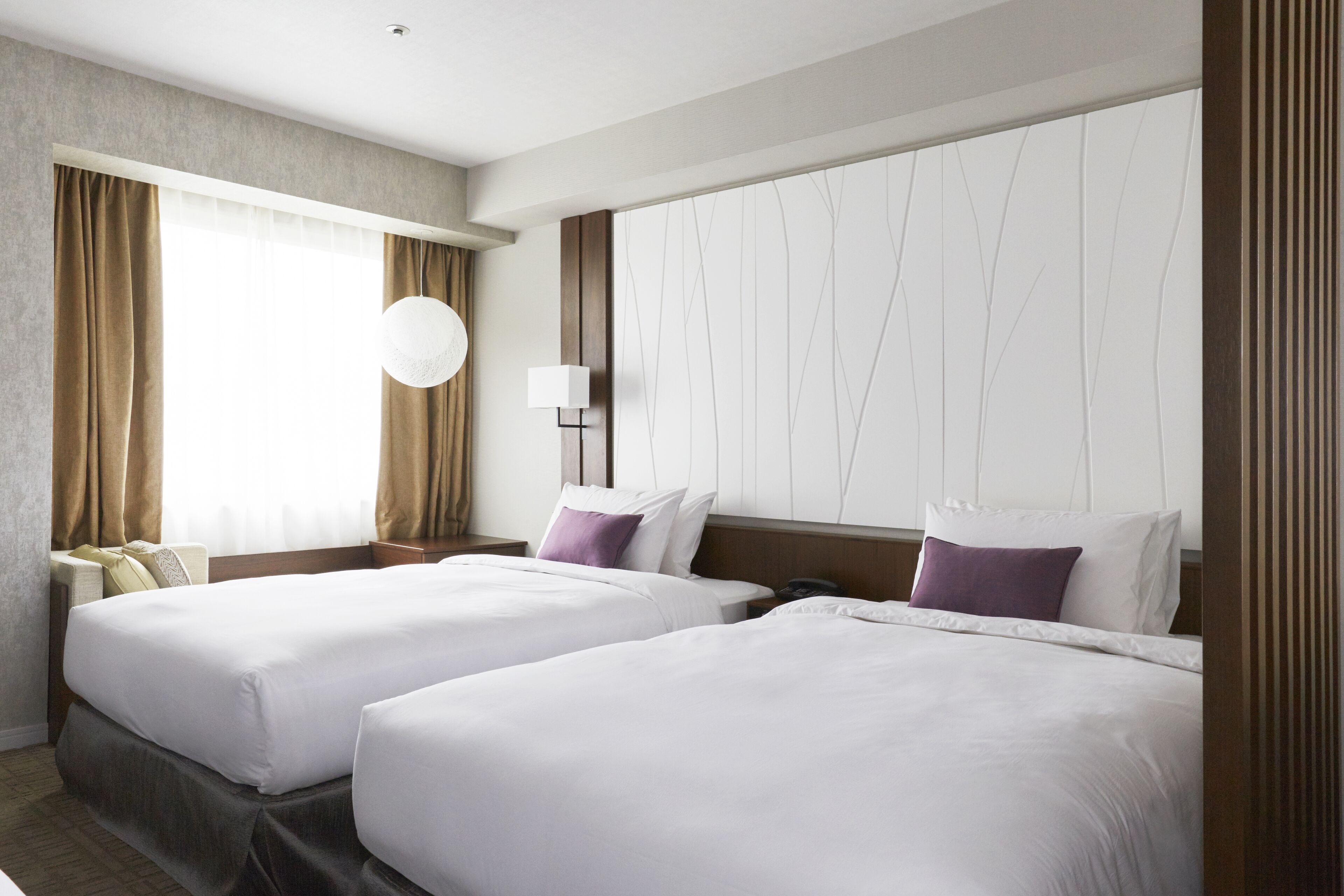 A hotel room with two beds, a large window with curtains, a wall-mounted lamp, and a round light fixture.