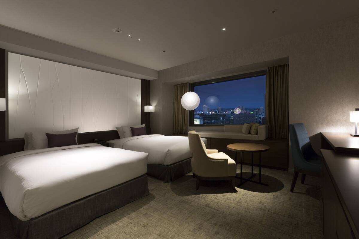 Ein Hotelzimmer mit zwei Betten, einer Couch, einem Schreibtisch und Blick auf die beleuchtete Stadt.