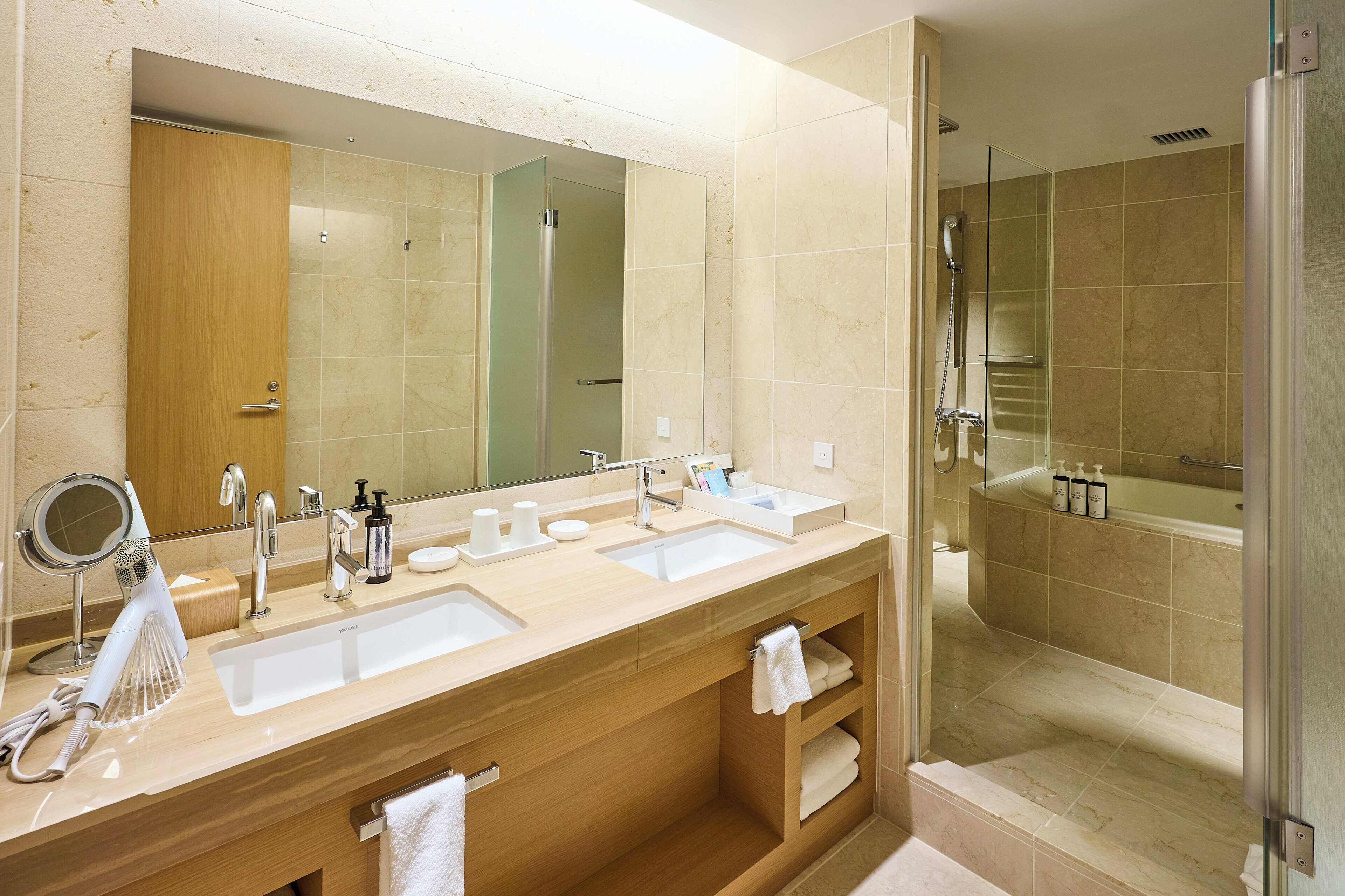 A modern bathroom with a large mirror, two sinks, a shower, and a vanity with a towel rack.