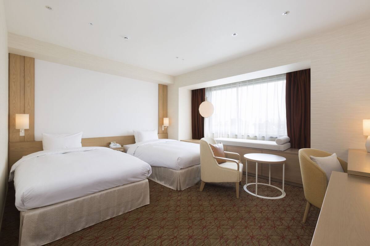 Ein Hotelzimmer mit zwei Betten, einem kleinen runden Tisch, zwei Sesseln, einer Couch und einem Fenster mit Vorhängen.