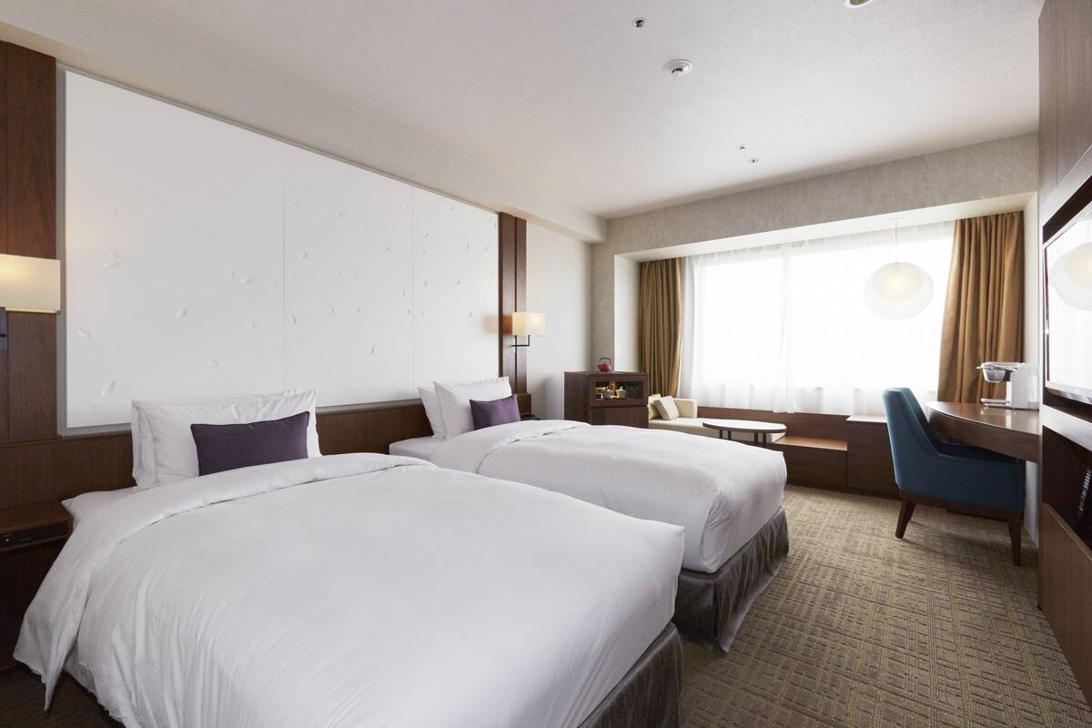 Ein Hotelzimmer mit zwei Betten, einem Schreibtisch, einem Stuhl, einem Fenster mit Vorhängen und einem an der Wand befestigten Fernseher.