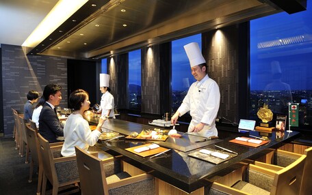 A modern restaurant with a chef preparing food at a counter. Guests are seated at tables with a view of the cityscape through large windows.