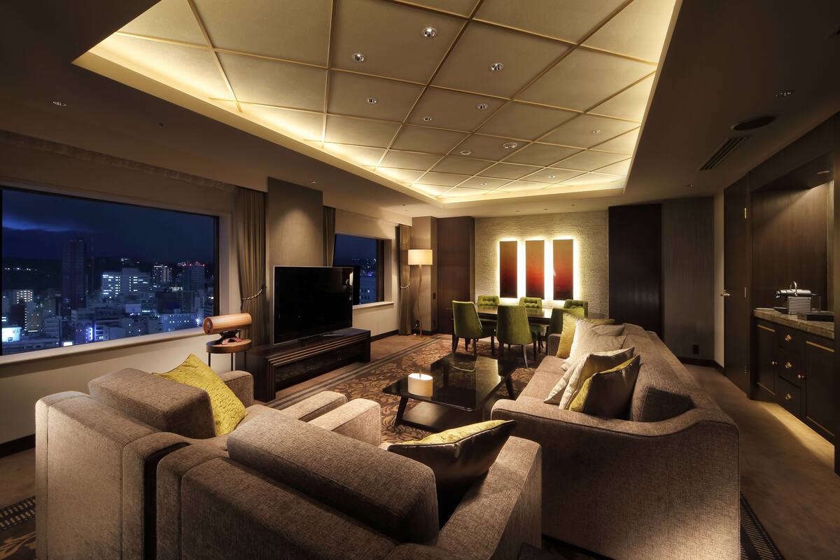 A modern hotel living room with a large TV, comfortable seating, a dining area, and a city view.