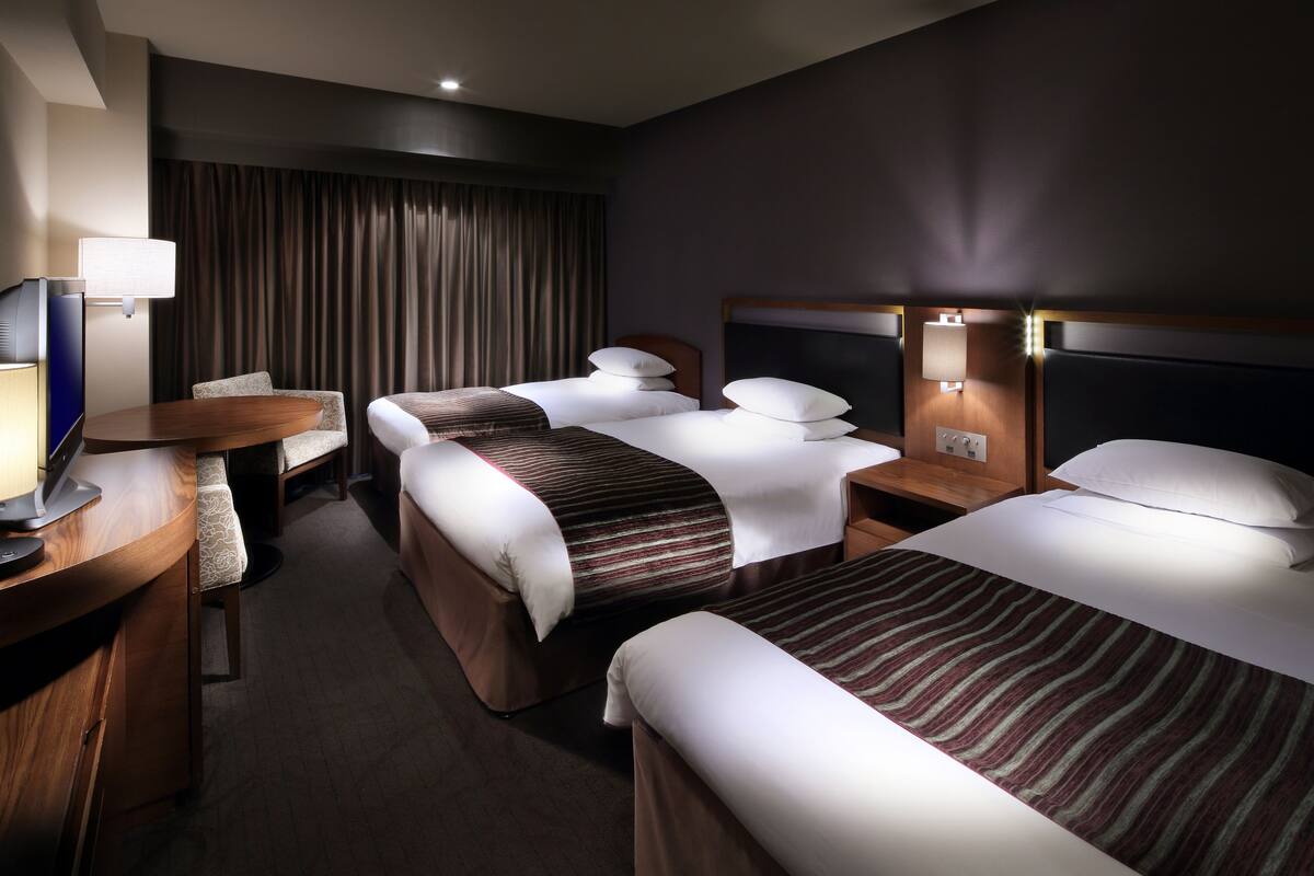 A hotel room with three beds, a desk with a lamp, and a TV.