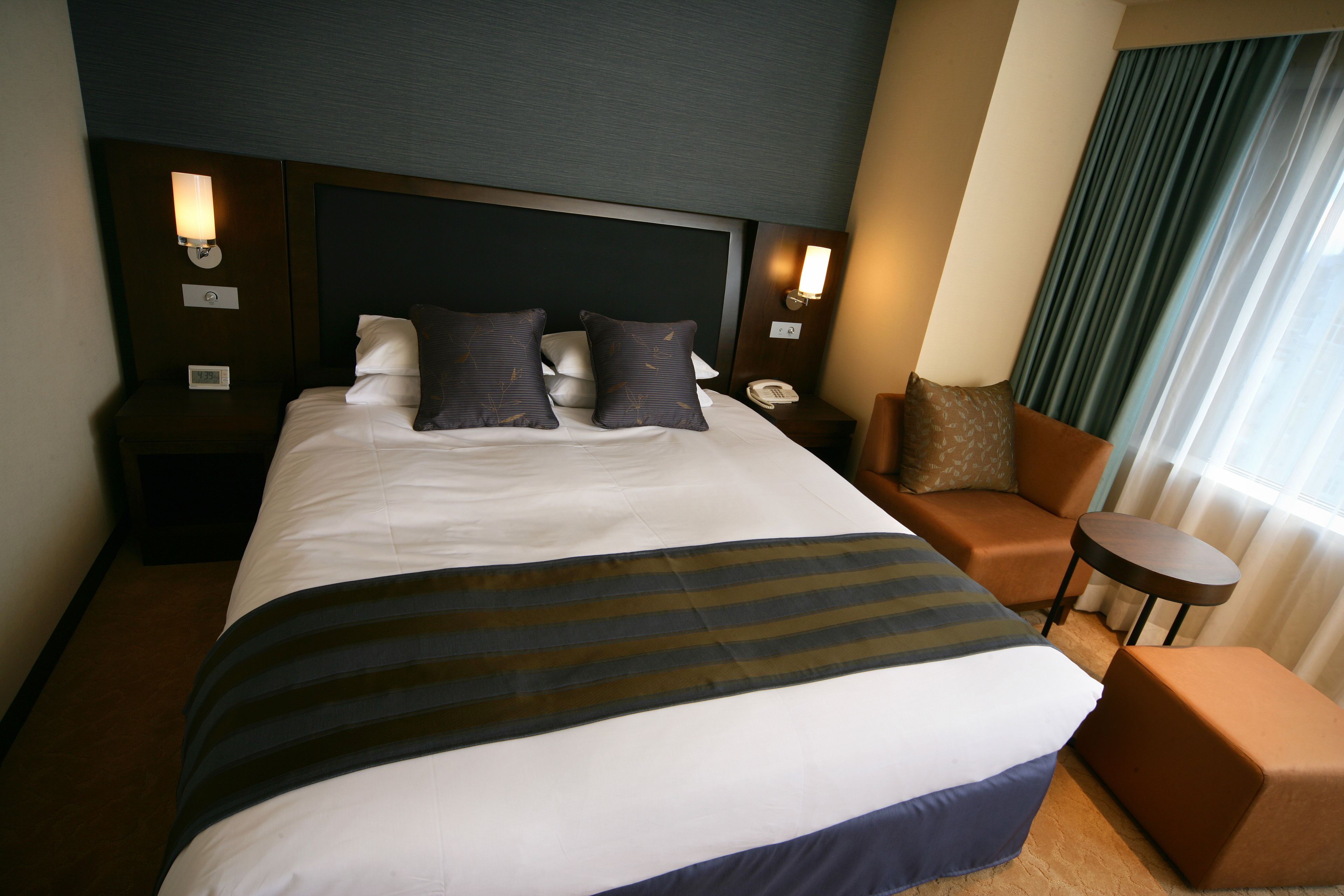 A hotel room with a large bed, two bedside tables with lamps, a sofa, a small round table, and a view of the outside through a window with curtains.