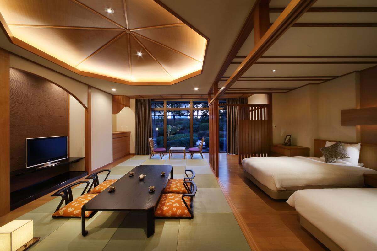 A modern hotel room with a dining table, two beds, a TV, and a view of the outdoors.