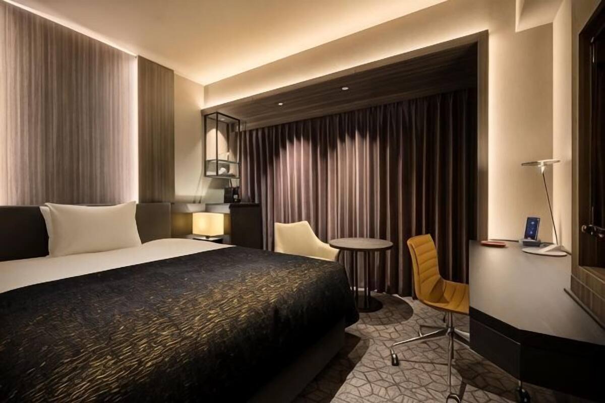 A hotel room with a large bed, a desk with a chair, a small round table, and a TV.