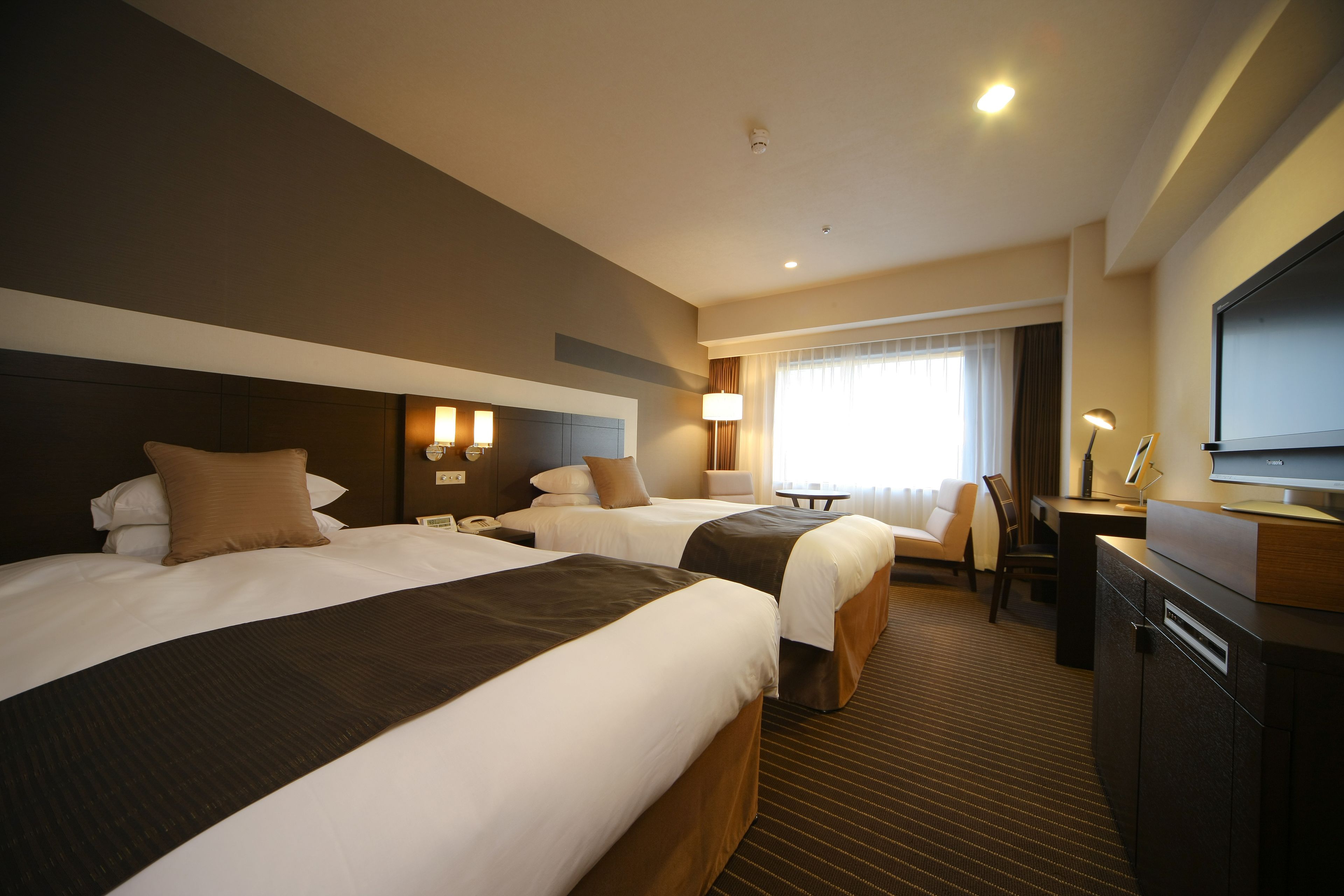 A hotel room with two beds, a desk, a television, and a window with curtains.