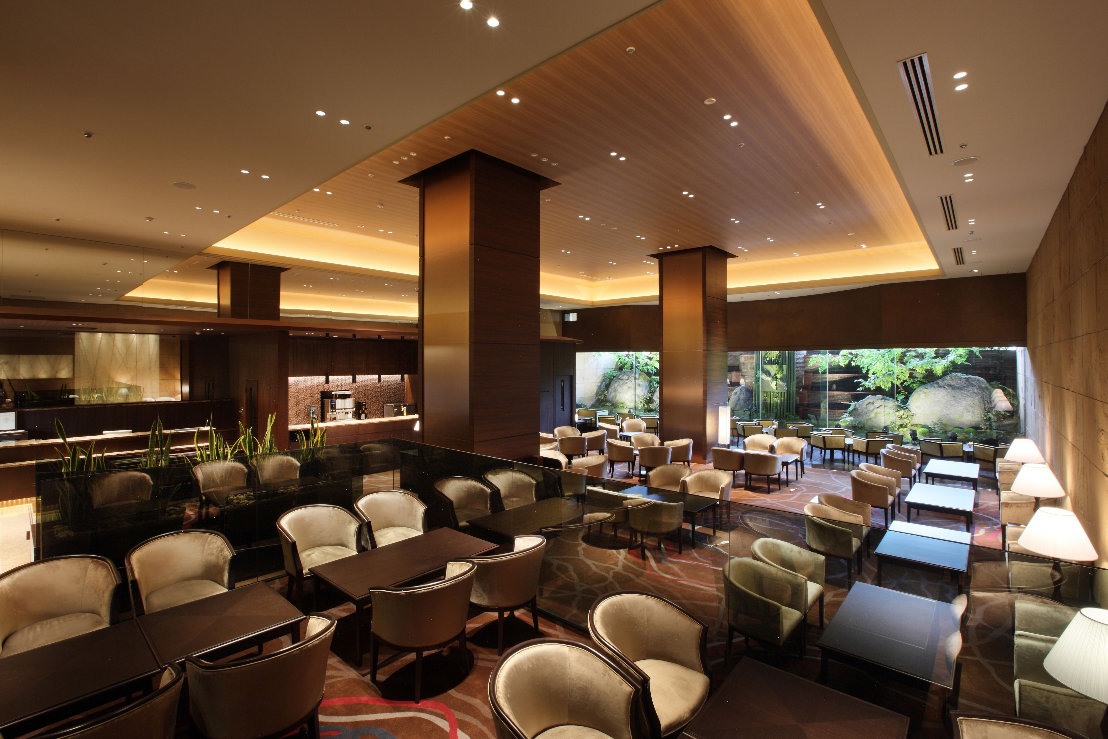 Un'ampia hall d'albergo con diverse zone salotto, un bar e un'ampia finestra che offre vista sul verde.