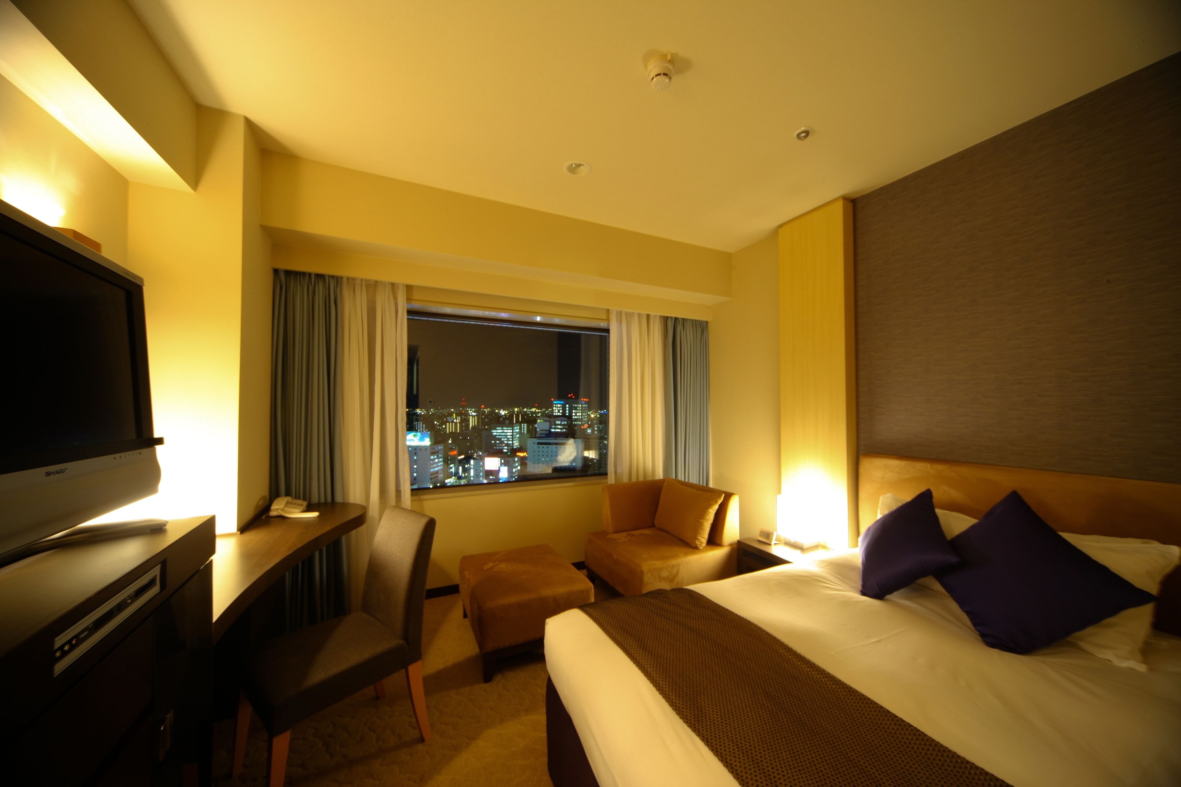 Une chambre d’hôtel avec un lit, un bureau avec une télévision, une chaise et une vue sur la ville la nuit.