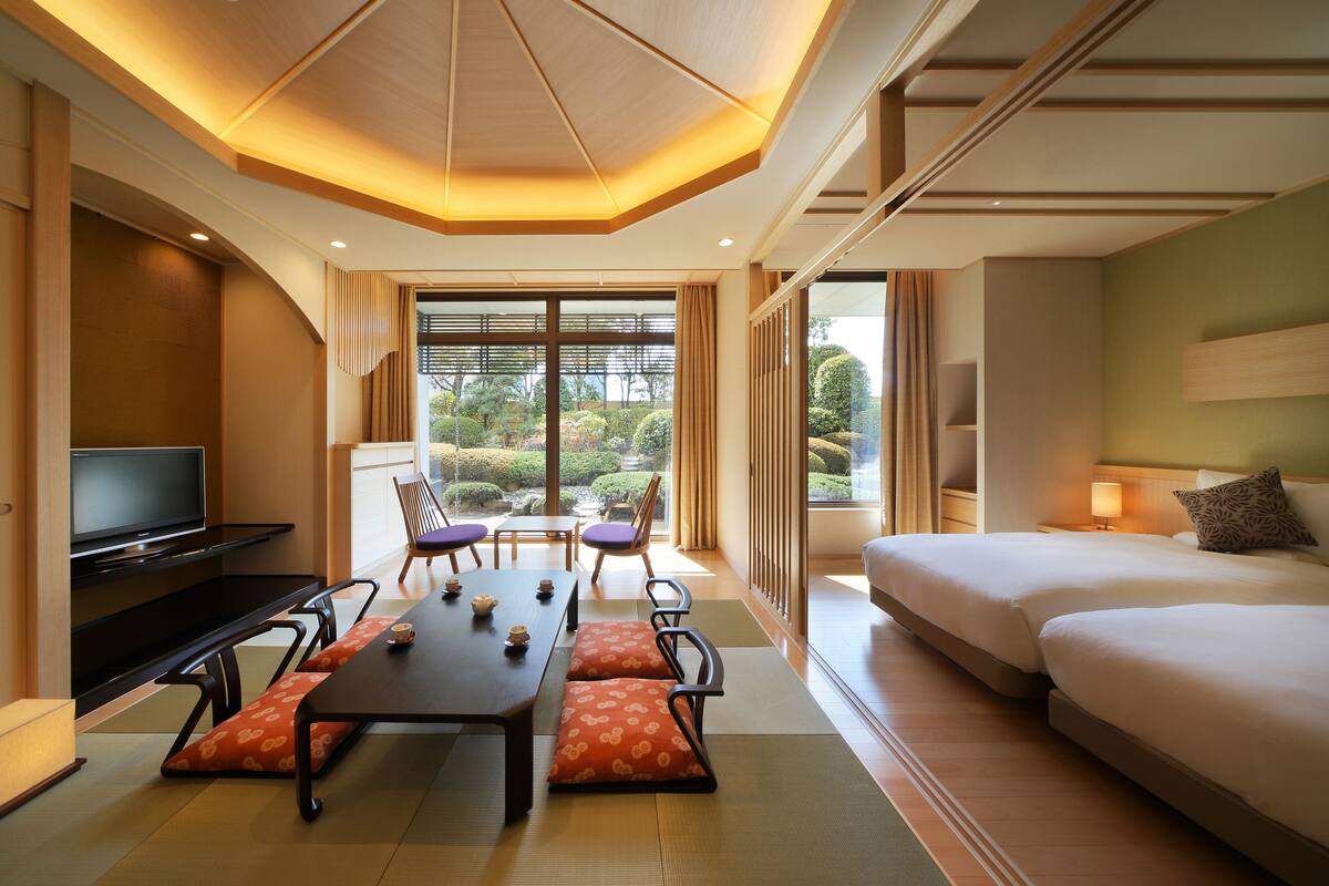 A hotel room with a large bed, a dining table with chairs, a TV, and a view of the garden.