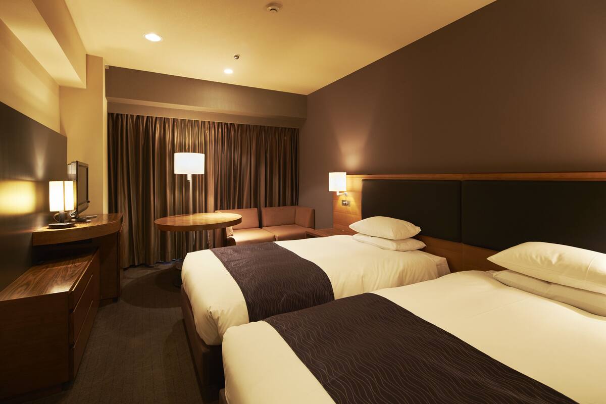 A hotel room with two beds, a desk, a chair, a round table, and a large window with curtains.