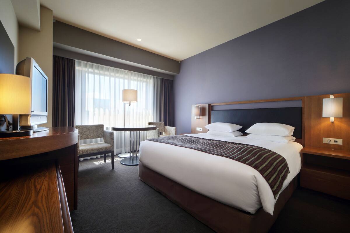 A hotel room with a large bed, a desk with a lamp, a chair, and a small table.