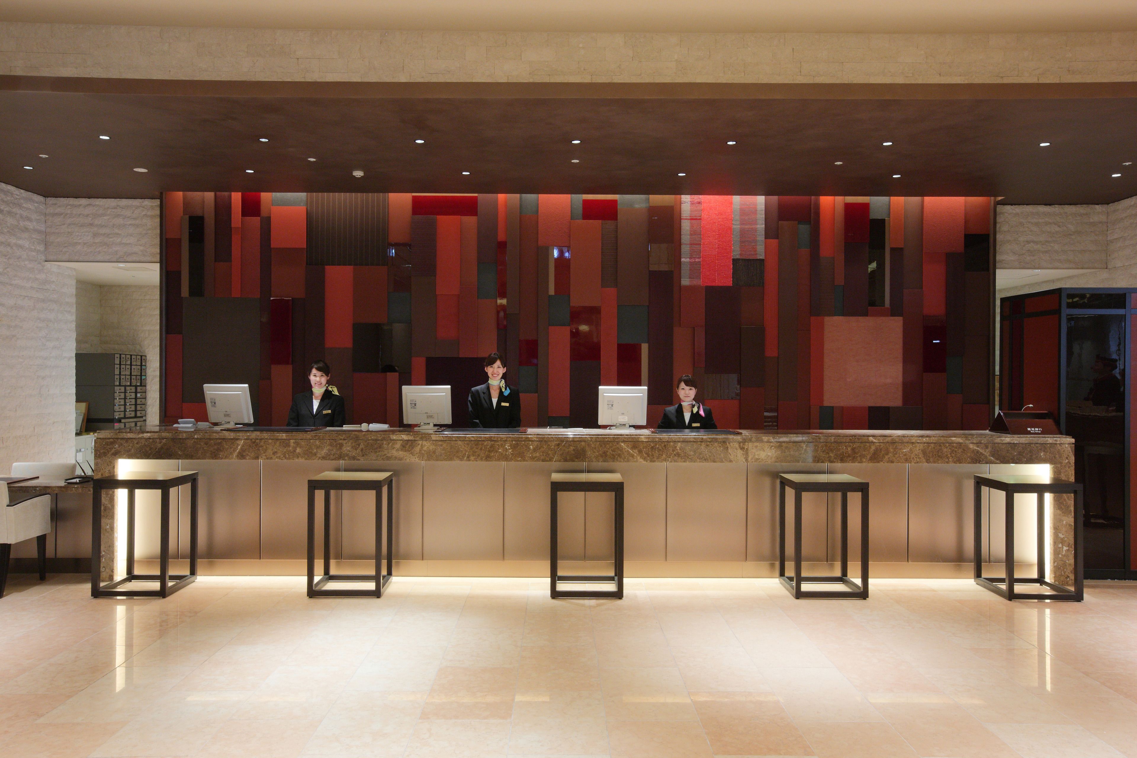 A hotel reception area with a modern design, featuring a reception desk, three staff members, and four bar stools.