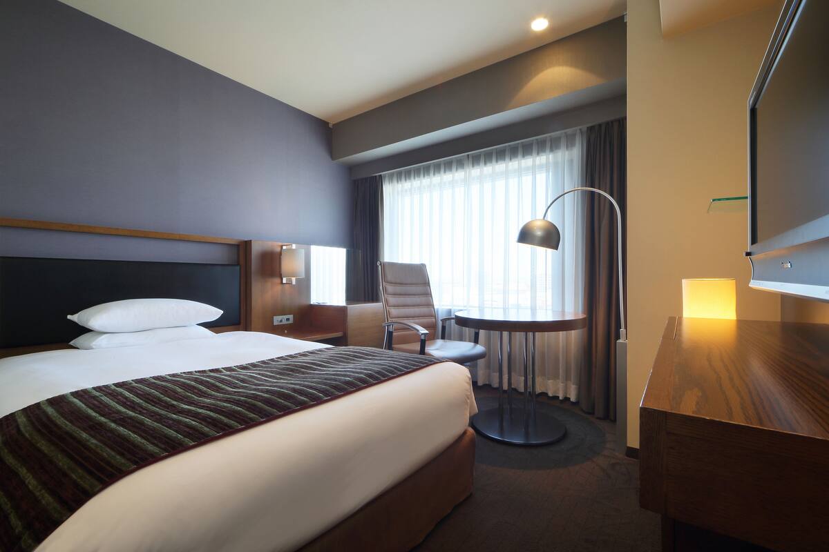 A hotel room with a large bed, a desk, a chair, a lamp, and a TV.