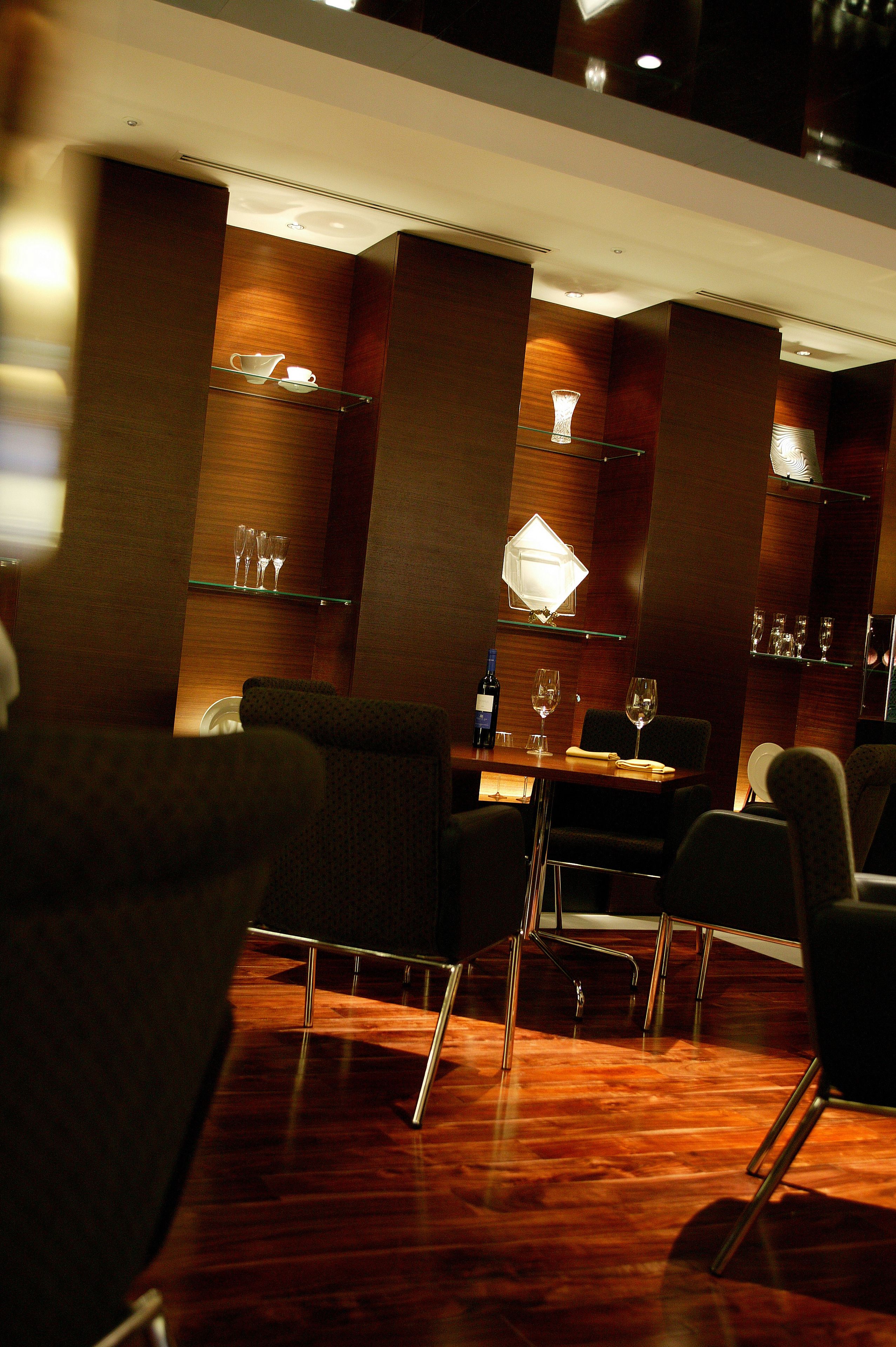 A modern interior with a wooden floor, a dining area with chairs and a table, and shelves with glasses and a bottle.