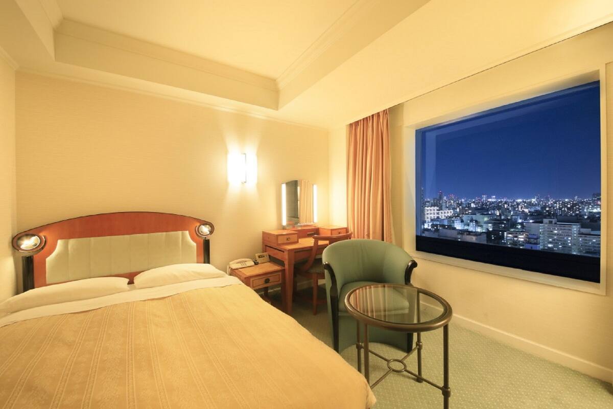 A hotel room with a bed, a desk, a chair, a small table, and a large window with a city view.