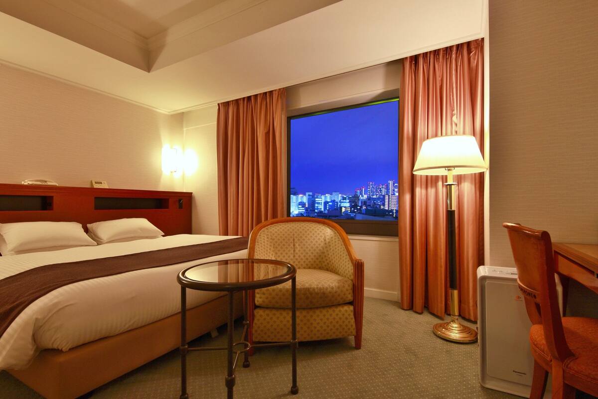 A hotel room with a bed, a chair, a table, a lamp, and a view of a cityscape at night.