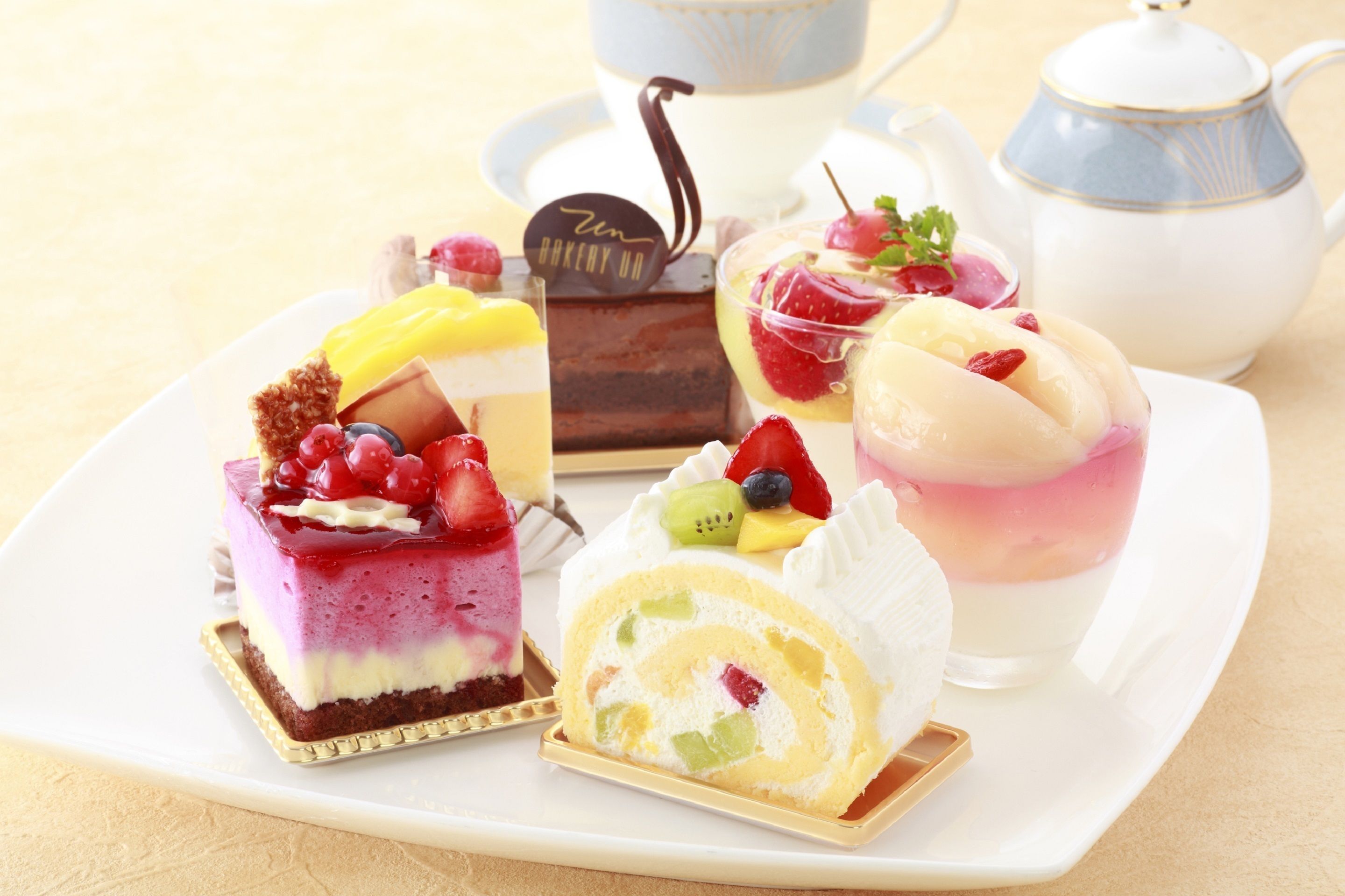 Una variedad de postres en una bandeja, incluyendo un pastel en capas, un bizcocho enrollado y un parfait, con una tetera y una taza al fondo.