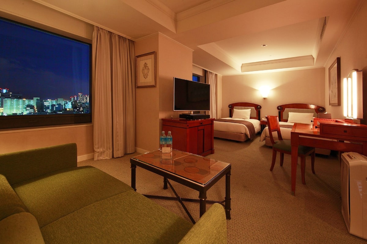 A hotel room with a large window, a television, a desk, a chair, and a bed.