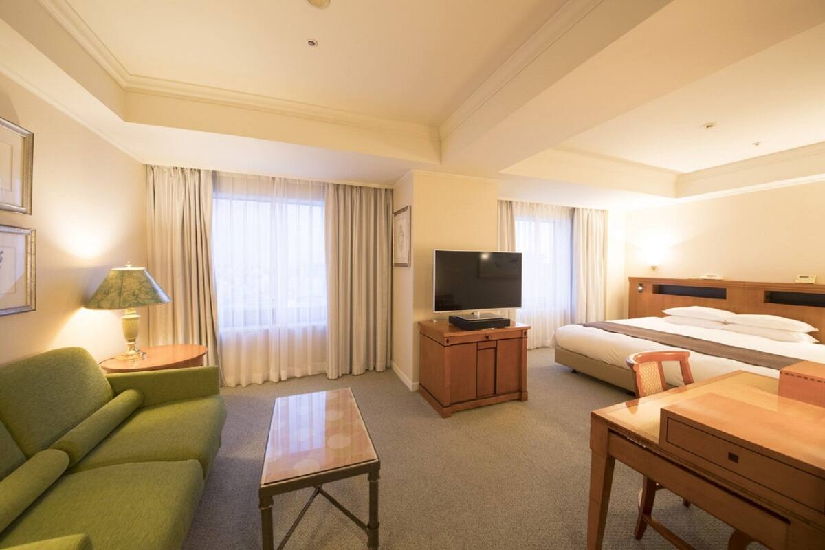 A hotel room with a green sofa, a wooden TV stand, a bed, a desk, and a lamp.