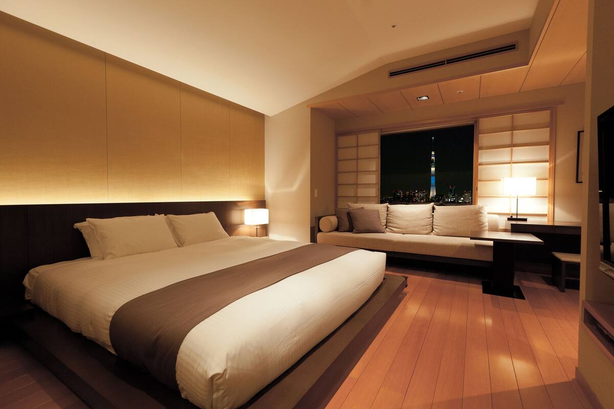 A modern hotel room with a large bed, a sofa, a desk, and a nightstand.