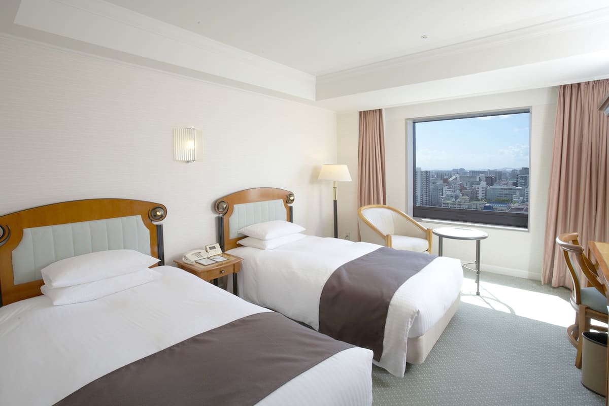 A hotel room with two beds, a desk, a chair, a lamp, and a window with a city view.