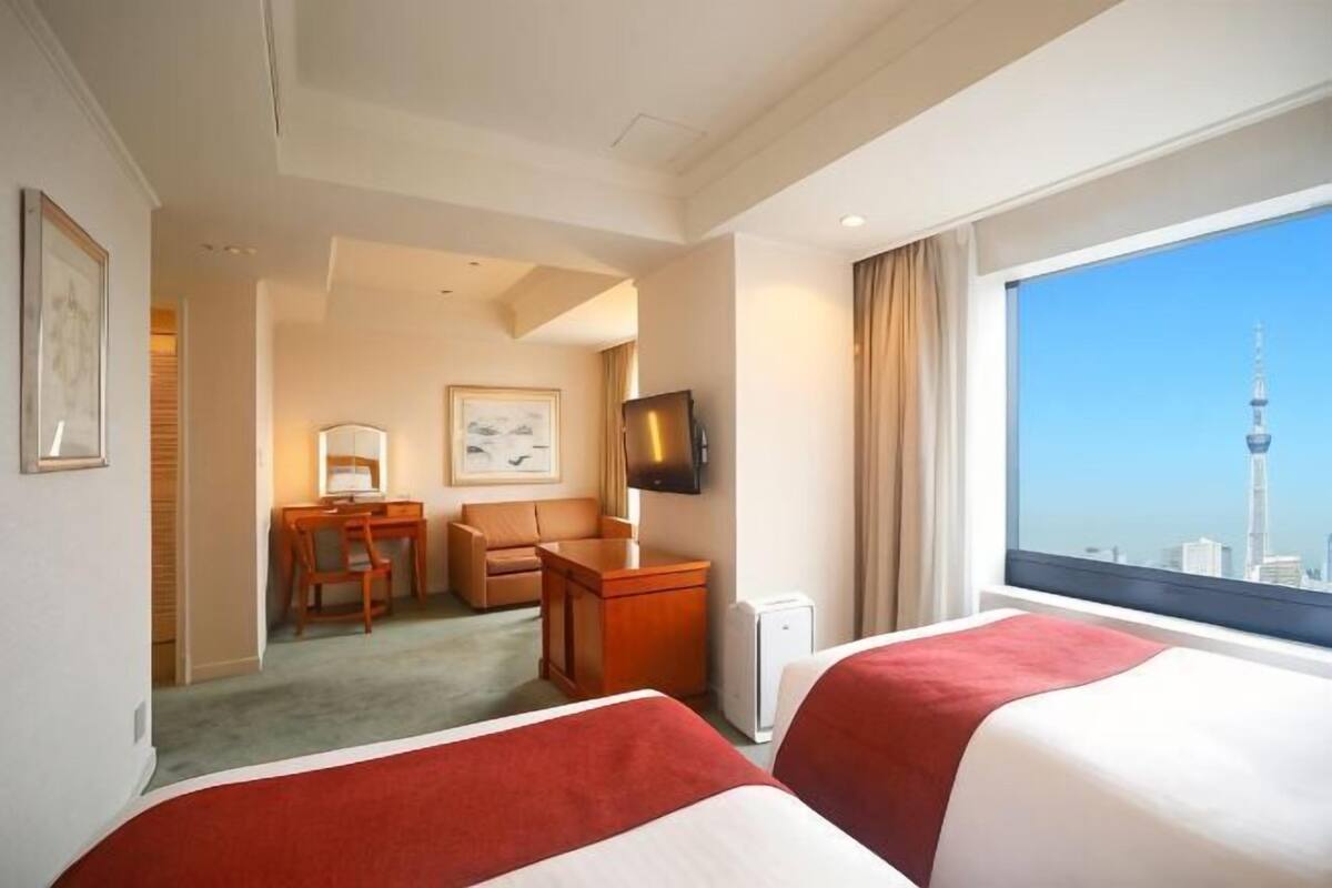 A hotel room with two beds, a desk, a chair, a television, and a large window with a cityscape view.