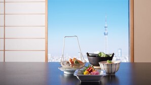 A table with a metal strainer basket, a black bowl with green vegetables, and a white bowl with colorful vegetables. A cityscape with a tower is visible through a window. - Hotel East 21 Tokyo (Tokyo)