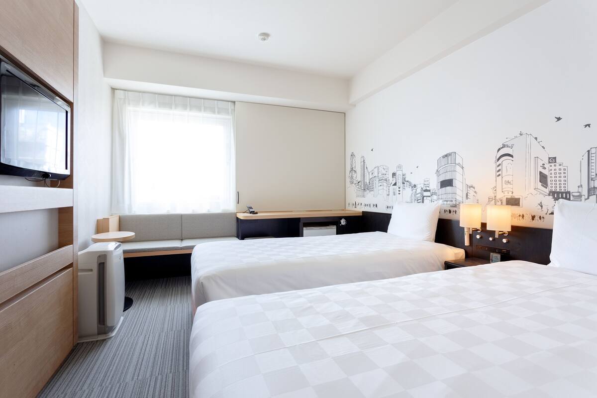 Ein Hotelzimmer mit zwei Betten, einem Schreibtisch, einem Sessel, einem Fernseher und einem Wandgemälde mit Stadtansicht.
