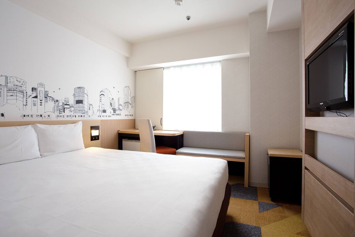 Ein Hotelzimmer mit einem großen Bett, einem Schreibtisch, einem Sessel, einem Fernseher und einem Fenster mit Jalousien.